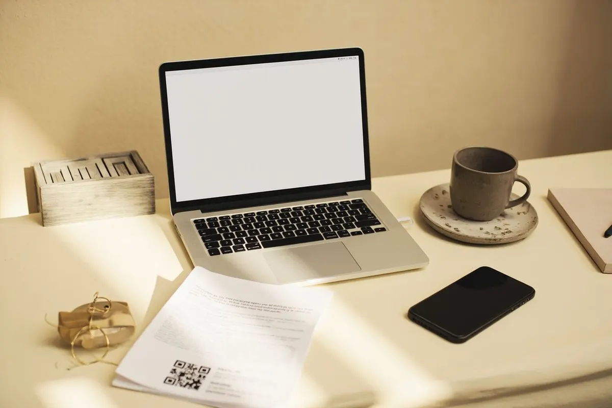 A laptop, a printed flyer with a QR code, a phone and a coffee cup on a sunny desk.