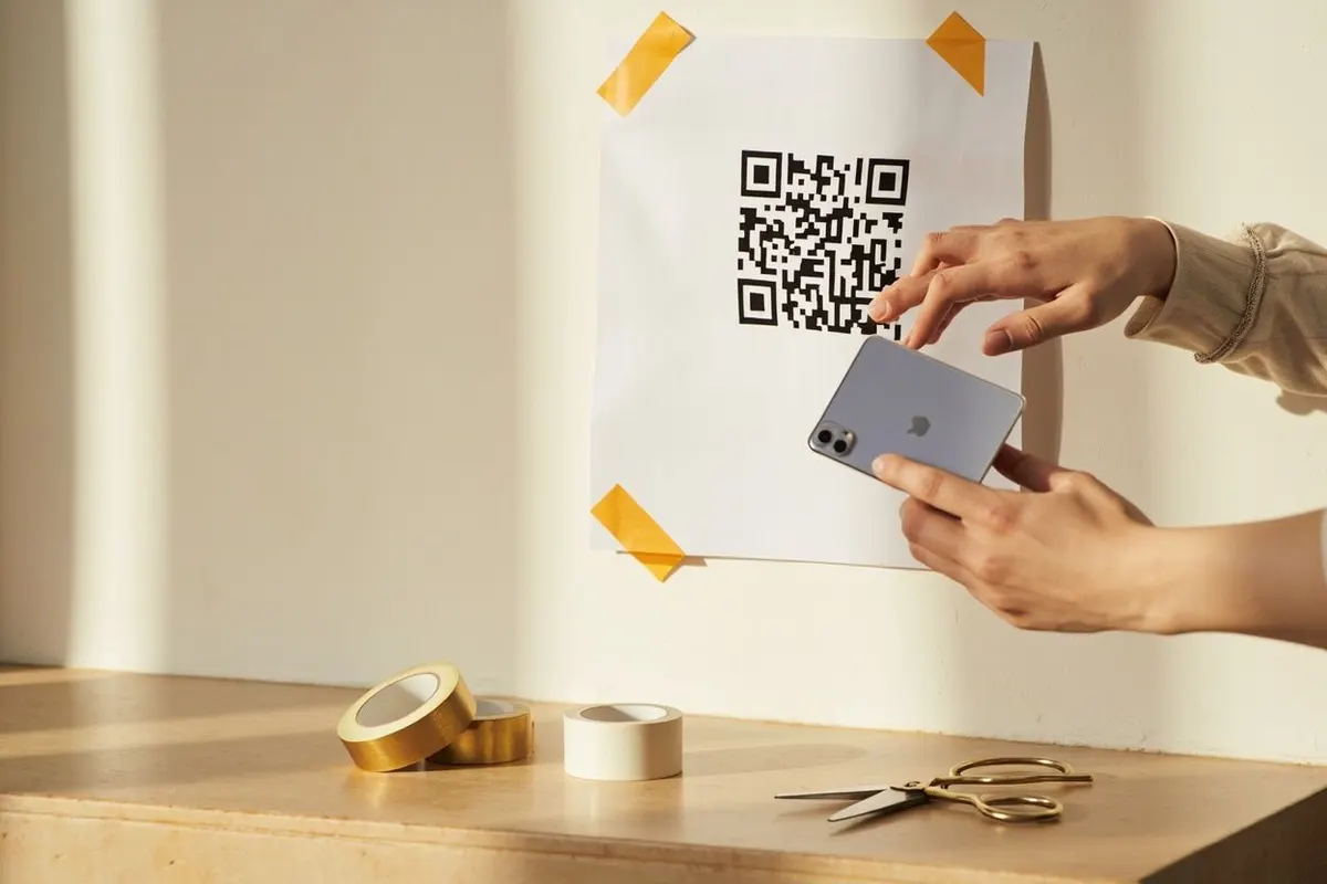 Hands scanning a QR code poster taped to a wall, with tape rolls and scissors on a shelf below.
